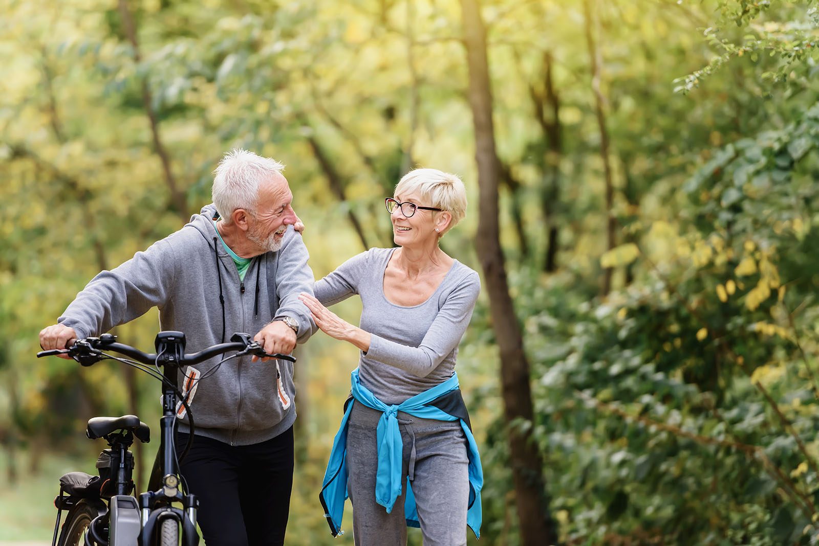 A smiling couple in their sixties walking their bicycles together through a sunny park